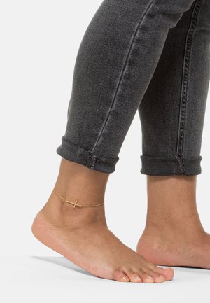 Zlatna anklet s križom na golom stopalu. Članak je ukrašen delikatnim dizajnom, upotpunjenim smrčenim sivim trapericama.