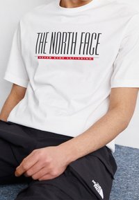 Bílý bavlněný tričko s logem "The North Face" v černém a červeném písmu, kulatým výstřihem, krátkým rukávem, nošené s černými kalhotami.