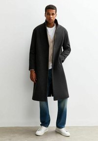 Cappotto di lana grigio scuro con collo alto, abbinato a un gilet beige con bottoni sopra una t-shirt bianca e jeans blu, completato da sneakers bianche.