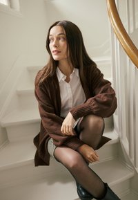 Cardigan en tricot marron porté sur une chemise blanche boutonnée, assis sur des escaliers blancs avec une rampe en bois. Collants foncés et bottes noires portés.