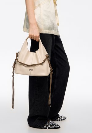 Persona con vaqueros anchos negros y una camiseta crema, sosteniendo un bolso beige con asa trenzada marrón y detalles plateados.