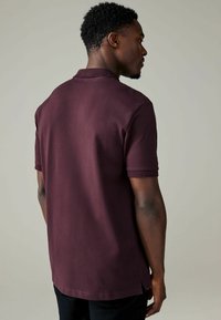 Polo tričko s krátkym rukávom v hlbokej burgundskej farbe, s golierom, postrannými rozparkami a hladkou textúrou, zobrazené zozadu.