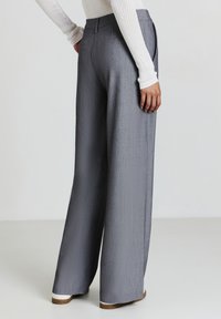 Persona che indossa pantaloni larghi grigi, un top bianco a costine a maniche lunghe e scarpe bianche, in piedi contro un fondo chiaro uniforme.