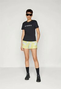 Άτομο που φοράει μαύρο μπλουζάκι Terrex, κίτρινες βερμούδες με σχέδια, μαύρες κάλτσες, μαύρα αθλητικά παπούτσια και σκούρα γυαλιά ηλίου, στέκεται μπροστά από μονόχρωμο φόντο.