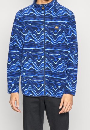 Mężczyzna ubrany w niebiesko-czarną, falowaną kurtkę zapinaną na zamek z małym wielobarwnym logo na klatce piersiowej, w zestawieniu z czarnymi spodniami.