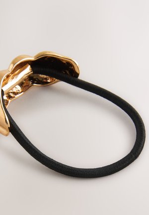 Gold-tone metal hair accessory with a wavy design attached to a black elastic band, featuring a shiny, smooth surface and textured interior.