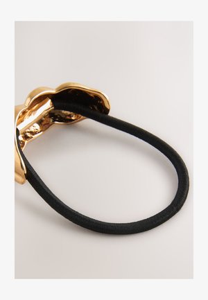 Gold-tone metal hair accessory with a wavy design attached to a black elastic band, featuring a shiny, smooth surface and textured interior.