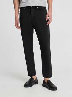 Uomo che indossa jeans neri slim fit e mocassini in pelle nera, in piedi contro uno sfondo chiaro uniforme.