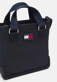 Marineblauw nylon tote bag met twee handvatten bovenaan, voorzien van een Tommy Jeans logo patch in rood, wit en blauw. Soepele textuur en gestructureerd ontwerp.