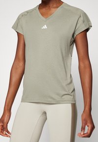 Persona vestida con una camiseta deportiva de cuello en V de manga corta en un verde oliva claro con un logo blanco y leggings ajustados beige, contra un fondo liso.