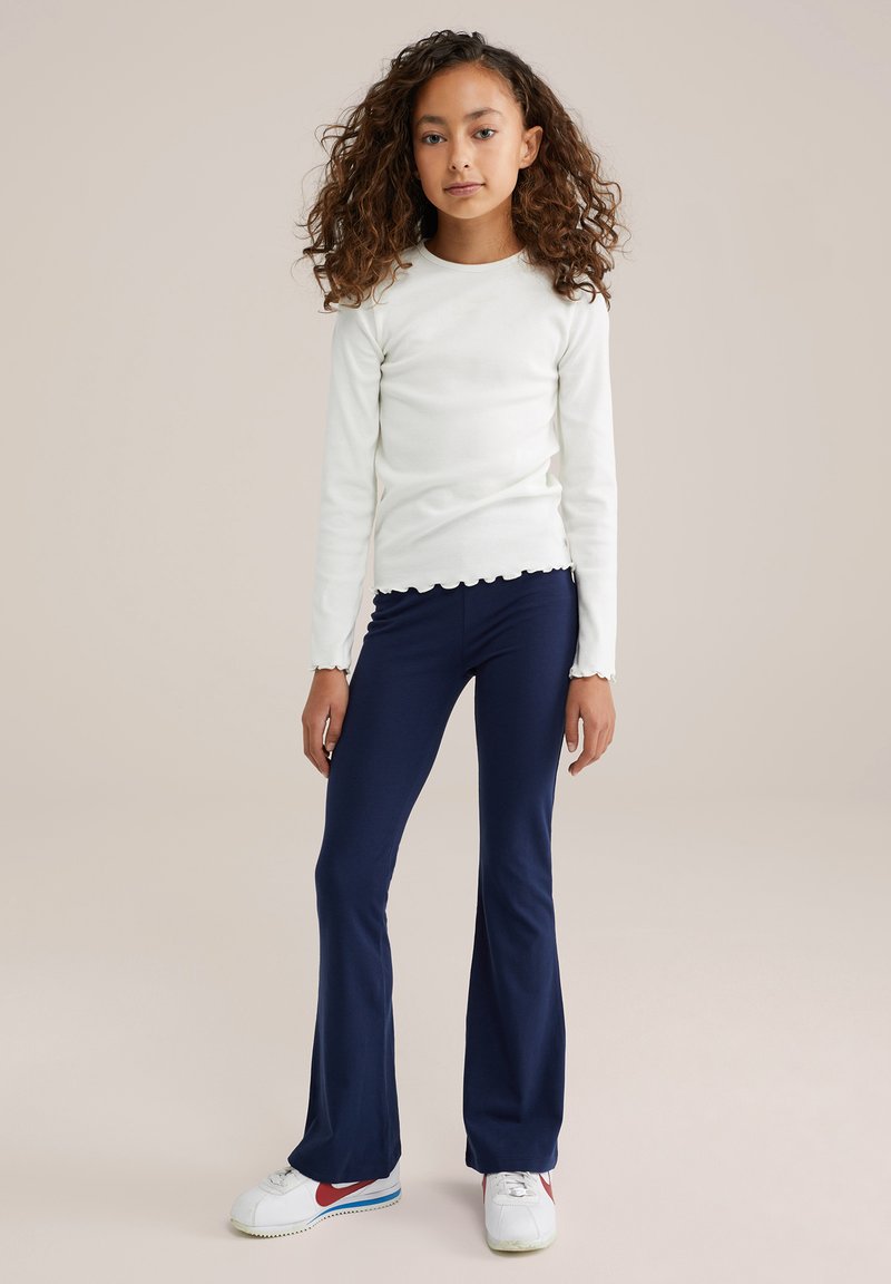 T-shirt blanc à manches longues avec un ourlet festonné, associé à un pantalon à pattes d'éléphant bleu marine, complété par des baskets blanches avec des accents rouges.