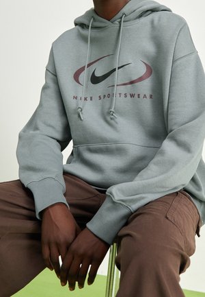 Personne portant un sweat à capuche Nike Sportswear vert tamisé avec cordons et un pantalon marron, assise avec les mains reposant entre les jambes.