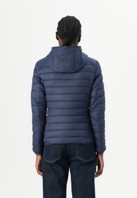 Person mit geflochtenen Haaren, die eine marineblaue gesteppte Daunenjacke mit Kapuze und dunkelblaue Jeans trägt, von hinten betrachtet.