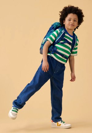 Enfant portant un t-shirt rayé vert et blanc, jean bleu, baskets blanches et sac à dos bleu foncé, debout sur une jambe sur fond beige.