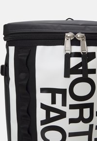 Biała torba izolacyjna z czarnym tekstem i akcentami, z zamknięciem na zamek błyskawiczny z dwoma metalowymi suwakami i czarnymi teksturowanymi paskami.