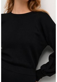 Femme portant un pull noir ajusté à manches longues avec un col rond, se tenant contre un mur blanc uni.