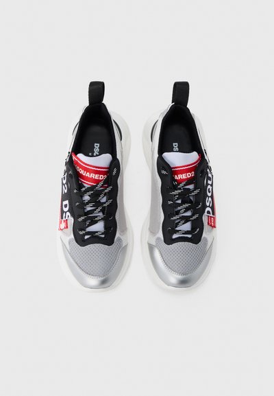 Τα αθλητικά παπούτσια διαθέτουν ασημί μεταλλική μύτη και τακούνι, μαύρα δίχτυ πλευρών, λευκά και μαύρα κορδόνια, και κόκκινη ετικέτα με το λογότυπο. Μινιμαλιστικός σχεδιασμός.