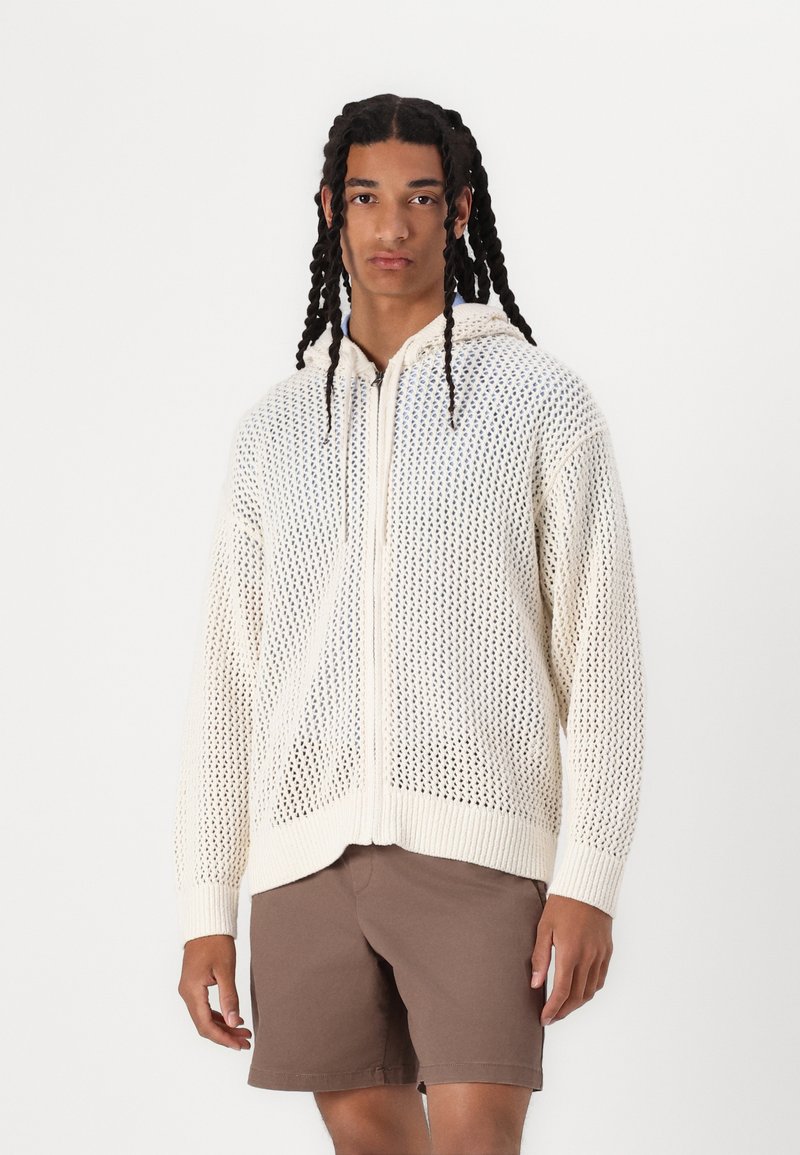 Felpa con zip in maglia color crema, con una texture simile a rete, dotata di cappuccio e polsini a costine. Indossata con pantaloni corti marroni.
