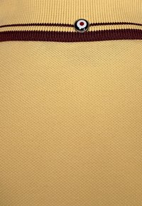 Tissu en tricot jaune avec deux rayures horizontales grenat et un petit bouton noir circulaire avec un motif cible rouge et blanc.