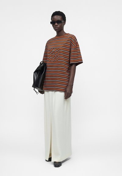 Άτομο που φοράει καφέ και μαύρο ριγέ φαρδιά πουκαμίσα, πλατιά λευκά παντελόνια με άνοιγμα μπροστά, μαύρα παπούτσια, γυαλιά ηλίου, κρατάει μαύρη τσάντα.