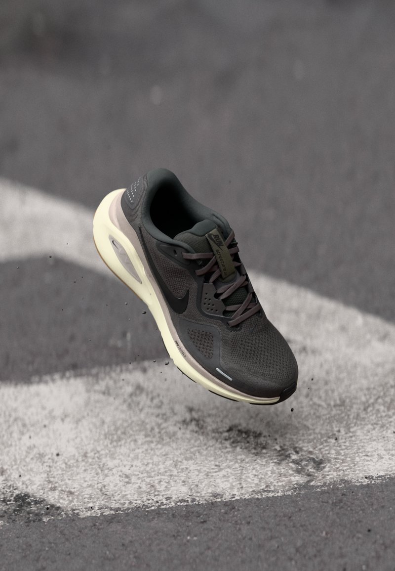Scarpa sportiva grigio scuro con suola bianca in volo sopra una strada asfaltata con segnaletica bianca e piccoli detriti.