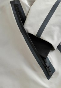Gros plan sur un tissu beige avec une fermeture éclair noire imperméable partiellement ouverte, montrant une doublure intérieure noire et des bandes réfléchissantes.