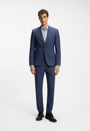 Jeune homme debout portant un costume bleu ajusté, une chemise à rayures et des chaussures noires habillées sur un fond clair uni.
