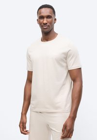 Άντρας που φοράει ανοιχτό μπεζ κοντομάνικη μπλούζα και παντελόνι στο ίδιο χρώμα, στέκεται μπροστά σε ένα απλό λευκό φόντο.