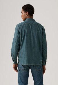 Camisa de ganga azul escura com mangas compridas, gola com botões e detalhes visíveis de costura, usada sobre jeans de ganga combinando.