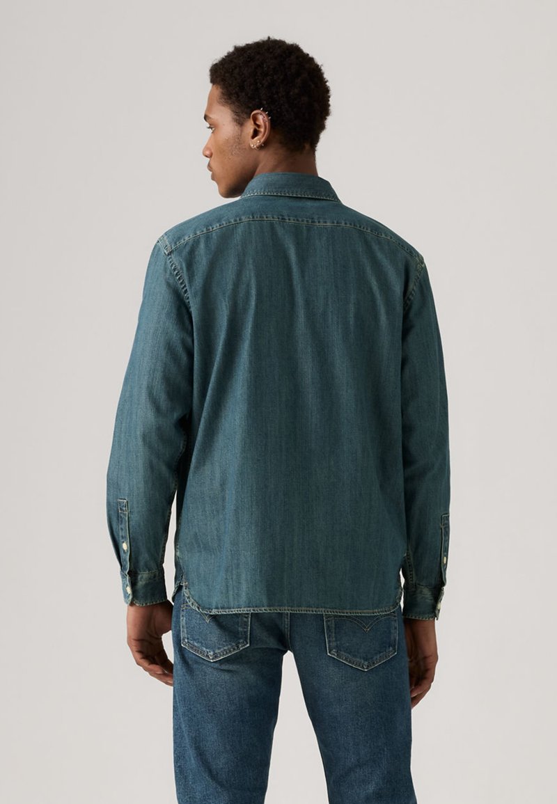 Camicia in denim blu scuro con maniche lunghe, colletto con bottoni e dettagli di cuciture visibili, indossata sopra jeans in denim abbinati.