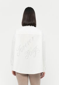 Camicia bianca con bottoni, caratterizzata da "Forever Hugo" decorato con strass sul retro. Maniche lunghe e orlo curvo aggiungono un tocco moderno.