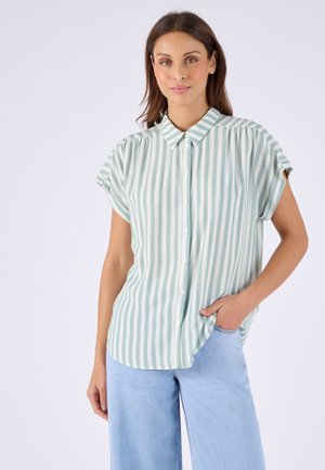 Femme portant une chemise boutonnée à manches courtes avec des rayures verticales vert clair et blanches, associée à un jean bleu clair, posant avec une main dans la poche.