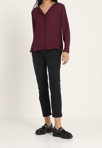 Blusa lunga marrone con colletto a girocollo, abbinata a pantaloni neri alla caviglia e scarpe nere con zeppa e dettagli con fibbia.