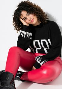 Maglione nero con la scritta "HAPPY" in bianco, maniche con texture; leggings rossi lucidi; stivaletti neri con dettagli borchiati. Sfondo bianco.