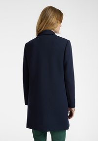 Abrigo de lana azul marino con cuello, mangas largas y corte recto. Presenta una textura suave y cae por debajo de las caderas. Diseño simple sin cierres visibles.