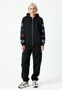 Felpa nera con zip e cappuccio, decorata con vari motivi sulle maniche, abbinata a pantaloni cargo neri e sneakers bianche. Tessuto liscio con vestibilità regolare.