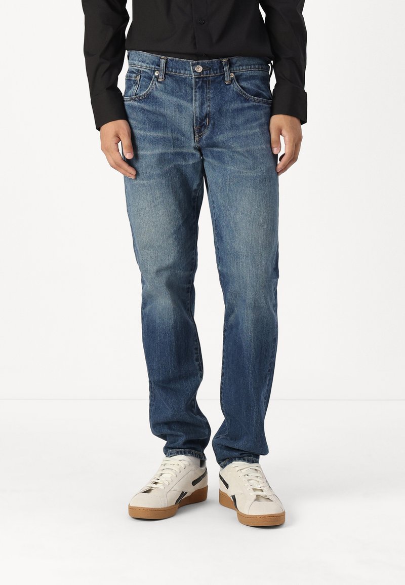Jeans de mezclilla azul con un corte recto, que cuentan con un cierre de cremallera, cinco bolsillos y un ligero desvanecimiento. Combinados con calzado blanco.