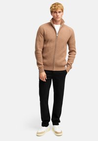 Beige ribbad zip-up tröja med hög krage, parat med svarta byxor och flerfärgade sneakers. Slät textur, figurnära design.