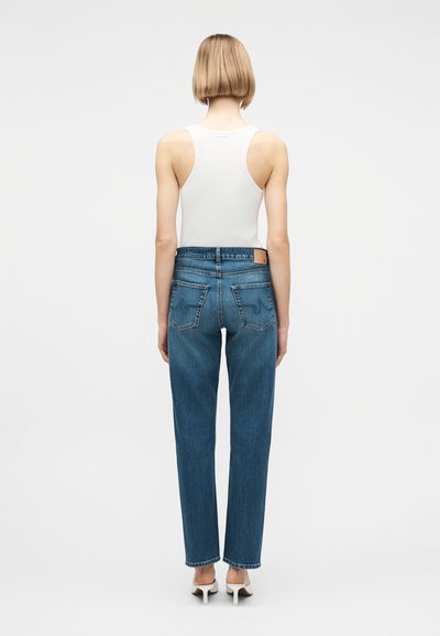 Γυναίκα με κοντά ξανθά μαλλιά που φοράει λευκό μπλουζάκι χωρίς μανίκια, ίσια μπλε τζιν παντελόνια και λευκά παπούτσια με ψηλό τακούνι, στέκεται γυρισμένη με την πλάτη.