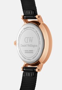 Rose gold stainless steel watch back with engraved branding and serial number. Black mesh strap visible at the bottom.