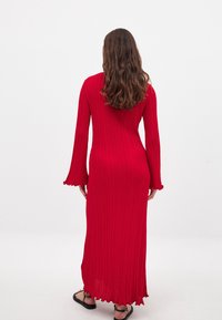 Lang rød ribbet kjole med flagrende ærmer og en let bølget kant, lavet af et let og tekstureret stof.