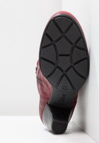 Botín de ante rojo con suela de goma negra texturizada. Tiene un tacón bajo, detalle de cremallera y superficie lisa. La suela presenta un agarre con patrón.