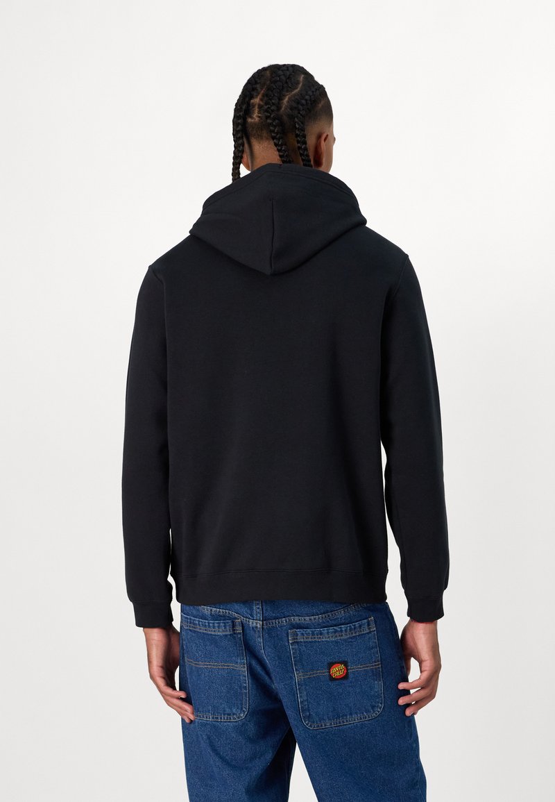 Sudadera negra con capucha ajustable, puños acanalados y bajo acanalado, combinada con jeans azules que presentan un parche de logo circular en el bolsillo trasero.