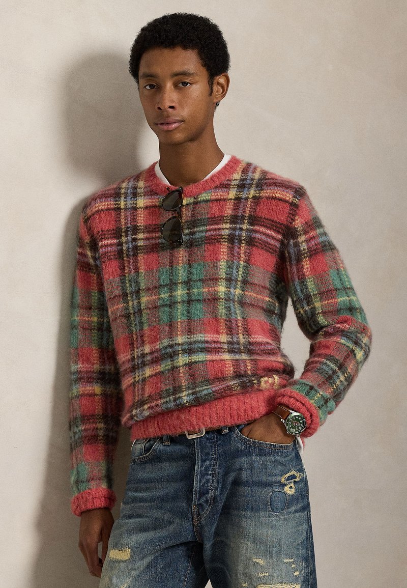 Stickad tröja i flerfärgat rutig mönster med rött, grönt och gult, med ribbade muddar och fåll. Bärs med slitna blå jeans.