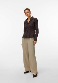 Chemise boutonnée marron foncé à manches longues, col avec revers, taille cintrée, associée à un pantalon large beige et des chaussures noires à bouts pointus.
