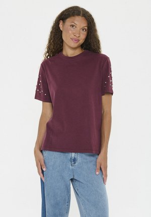Woman with curly hair wearing a burgundy short-sleeve shirt with pearl details on sleeves and light blue jeans, standing against white background.