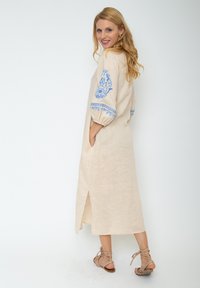 Beige Leinenkleid mit blauer floralem Stickerei auf den gepufften Ärmeln, seitlichen Taschen und einem Schlitz vorne. Kombiniert mit hellbraunen Sandalen.