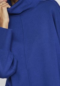 Maglione blu lavorato a maglia con collo a scialle, tessitura a coste e spalle scese. Presenta una cucitura verticale al centro. Design morbido e casual.