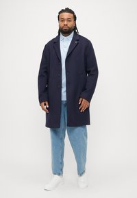 Cappotto di lana blu navy con tre bottoni neri, rever a incavo, tasche laterali, indossato sopra una polo azzurro chiaro e jeans azzurri larghi. Scarpe da ginnastica bianche.