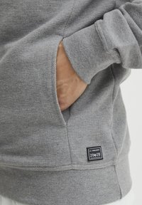 Grijze sweatshirt met textuurstof, zijinsneden en een zwart label met "20/20" branding op de onderste zoom.